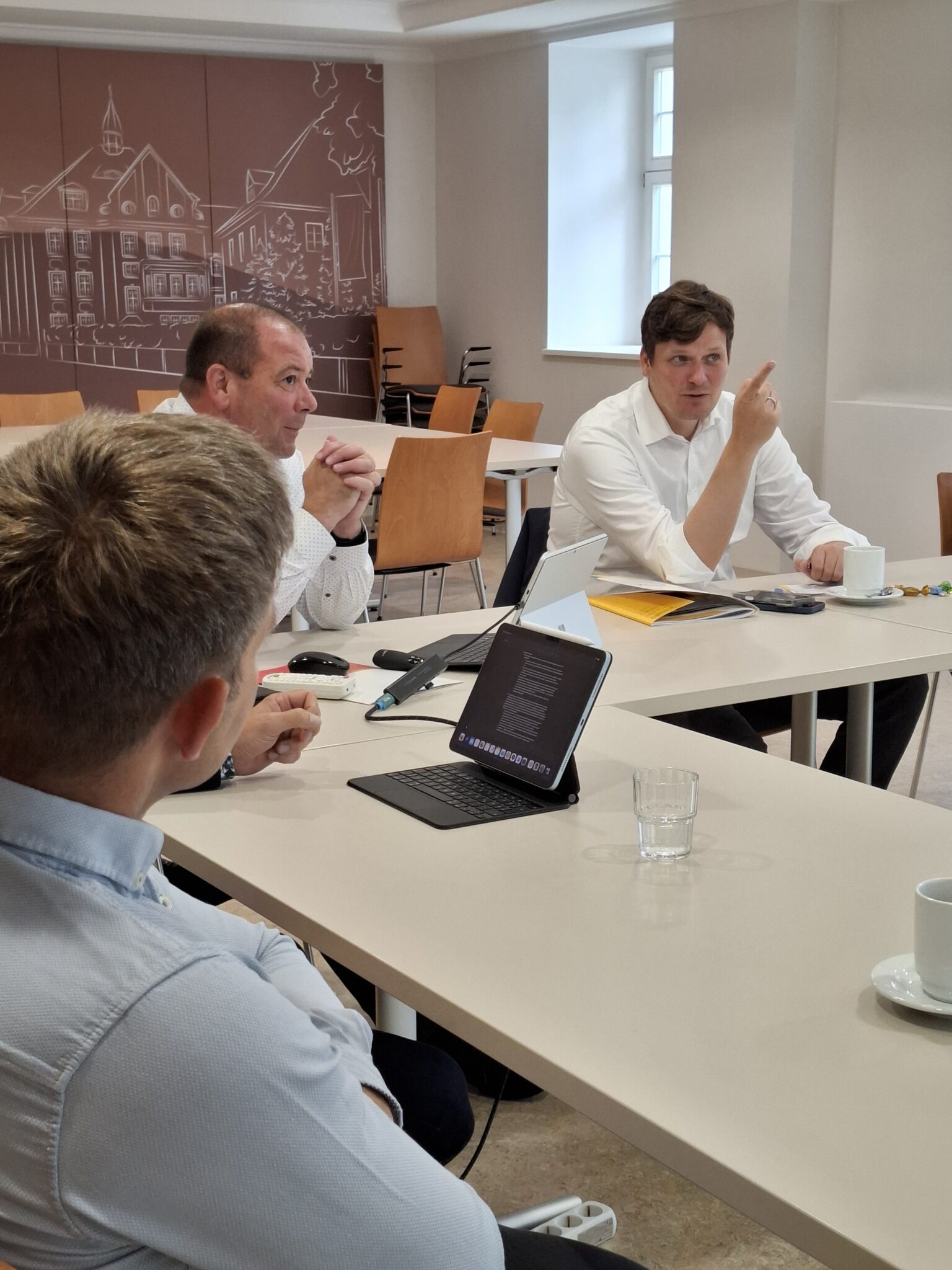 Der Kultusminister sitzt in einem weißen Hemd mit zwei Männern am Tisch und deutet mit dem Zeigefinger auf die Präsentation.