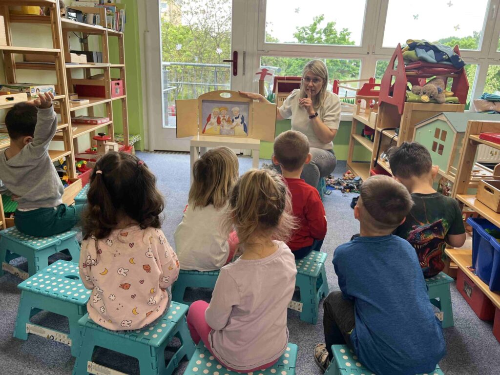 Erzieherin sitzt vor Kindern an einem Puppentheater und erzählt eine Geschichte während Kinder gespannt zuhören.