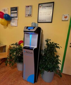 Wasserspender steht betriebsbereit an der Wand. Daneben stehen zwei Topfpflanzen.