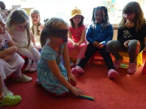 Kinder sitzen im Kreis und spielen das Speil "Blinde Kuh". Ein Kind sitzt in der Mitte und hat eine Augenbinde um.