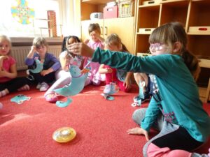 Kinder sitzen auf dem roten Teppich eines Zimmers im Kreis. Ein Mädchen in der Mitte der Kinder hält etwas gebasteltes nach oben.